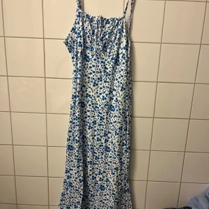 Vit långklänning med blå blommor - Säljer en vit långklänning med blått blommigt mönster och smala axelband. Klänningen har en avslappnad passform och knytning fram vid bysten. Perfekt för varma dagar och har en lätt, flowy känsla.