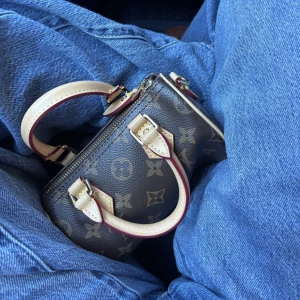 Louis Vuitton Mini Monogram Handväska - Säljer en ikonisk handväska från Louis Vuitton i klassiskt brunt monogrammönster. Väskan har rund form, två korta handtag i ljust skinn och guldfärgade metalldetaljer. Lite av guldfärgen har tyvärr nöts bort från dragkedjan och vissa delar men inget som märks. Det är en Nano Speedy med ett axelband. Jag köpte den från Sellpy innan för 6000kr därför säljer jag den för 4600 men pris går att diskutera.
