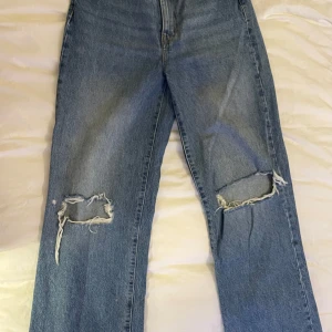 Blå ripped jeans från 157, storlek S - Säljer ett par blå jeans från 157 i storlek S. Jeansen har hög midja och raka ben med stora slitningar över knäna för en trendig, avslappnad look. Klassisk femficksmodell i denim med dragkedja och knapp framtill.