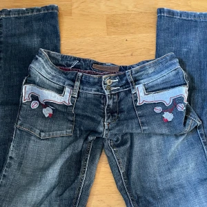 Blå lågmidjade jeans med broderade detaljer - Snygga blå jeans från Miss Sixty med unika broderade blommor och mönster på både fram- och bakfickor. Jeansen har dubbla knappar i midjan och coola kontrastsömmar. Perfekt för dig som vill sticka ut med detaljer och stil.