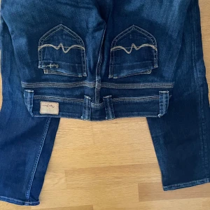 Mörkblå straight jeans från Pepe Jeans - Snygga mörkblå jeans från Pepe Jeans med raka ben och låg midja. Klassiska kontrastsömmar, coola bakfickor med broderad detalj och logopatch i läder baktill. Regular fit och tillverkade i bomull med en liten del elastan för stretch.