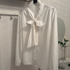 Vit blus med stor rosett i halsen - Säljer en vit blus med lång ärm och en stor rosett i halsen. Blusen har knappar framtill och är tillverkad i ett mjukt, lätt material som ger ett elegant intryck. Perfekt för dig som gillar klassisk och stilren look.