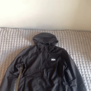 Svart vindjacka från Helly Hansen - Svart vindjacka från Helly Hansen med huva och vitt HH-logga på bröstet. Jackan har dragkedja framtill och meshfoder på insidan för extra komfort. Perfekt för blåsiga dagar och enkel att matcha med olika outfits.