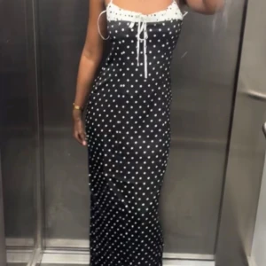 Svart lång prickig klänning med snörning - Supersnygg lång svart klänning med vita prickar och smala axelband. Överdelen har en vit kant med snörning framtill som ger en söt touch. Klänningen har en figurnära passform och är perfekt för dig som gillar en klassisk men trendig look.
