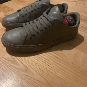 Grå sneakers från Axel Arigato - Gråa sneakers från Axel Arigato med stilren design och platt sula. Skorna är i skinn med matchande grå snörning och diskret logga bak på hälen. Perfekta för dig som gillar minimalistisk och clean stil.