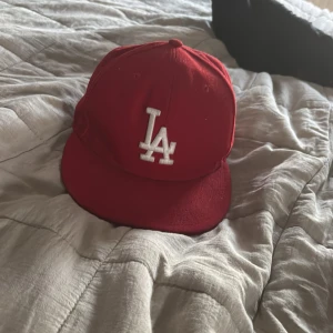 59fifty keps  - Säljer en röd keps från New Era med LA-logga broderad i vitt framtill. Klassisk 59FIFTY-modell med platt skärm och svart insida. Perfekt för dig som vill ha en streetig och sportig look.