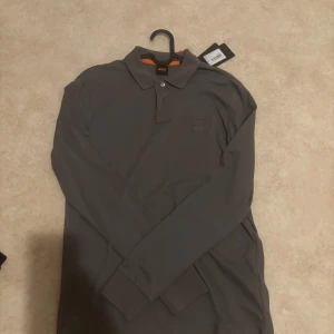 Grön långärmad pikétröja från BOSS - Snygg grön långärmad pikétröja från BOSS i slim fit-modell. Klassisk krage med knappar och diskret BOSS-logga på bröstet. Tröjan har ribbade muddar vid ärmsluten och är tillverkad i mjuk bomull för en clean och stilren look. Ny med prislapp på