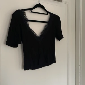 Svart croppad topp med spetsdetalj - Säljer en svart croppad topp med korta ärmar och djup v-ringning. Toppen har ribbad struktur och är dekorerad med spets längs urringningen för en extra snygg touch. Perfekt att styla med jeans eller kjol för en trendig look. 🍸