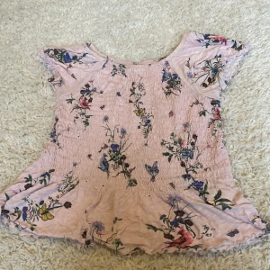 Blommig rosa blus från Lindex - Supersöt blus från Lindex i ljusrosa med blommigt mönster i flera färger. Modellen har korta ärmar, smockad överdel och lätt utställd nederkant. Perfekt för dig som gillar romantisk och färgglad stil. 40kr med frakt!📦 