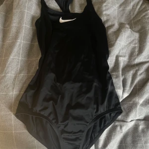 Svart baddräkt från Nike - Säljer en svart sportig baddräkt från Nike med klassisk vit logga framtill. Modellen har brottarrygg och öppen rygg för extra rörelsefrihet. Perfekt för simträning eller strandhäng. Materialet är stretchigt och snabbtorkande syntet.