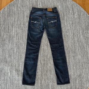 Nudie Jeans blå jeans W29 L32 - Säljer ett par mörkblå jeans från Nudie Jeans med klassisk femficksdesign och snygga kontrastsömmar. Jeansen har raka ben och coola broderade bakfickor med vågigt mönster. Tillverkade i slitstarkt denim av bomull. Perfekta för dig som gillar stilrena och tidlösa jeans.