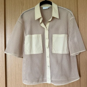 Beige nätskjorta från Coster Copenhagen  - Säljer en beige nätskjorta från Coster Copenhagen med kort ärm, två stora bröstfickor och tryckknappar framtill. Skjortan är luftig och har ett coolt nätmönster, perfekt för lager-på-lager. Oversized fit och riktigt trendig look.