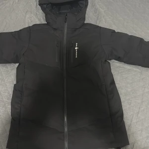 Svart pufferjacka från Sail Racing - Svart pufferjacka från Sail Racing med huva och två bröstfickor med dragkedja. Jackan har en clean look med logga på bröstet och hel dragkedja framtill. Perfekt för kalla dagar och riktigt snygg till streetwear-stilen.