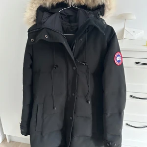 Svart dunjacka från Canada Goose - Säljer en svart Canada Goose dunjacka med pälskantad huva och klassisk logga på ärmen. Jackan har dragkedja, knappar och snörning vid huvan. Perfekt för kalla vinterdagar och har en stilren, lång modell.