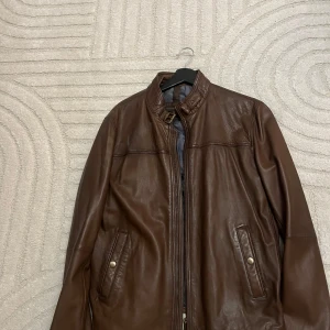 Brun skinnjacka med ståkrage - Snygg brun skinnjacka med ståkrage och dragkedja framtill. Jackan har två fickor med tryckknappar och stilrena sömdetaljer över bröstet. Perfekt för dig som vill ha en klassisk och tidlös look.