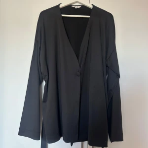 Svart omlottkofta med knytband - Svart omlottkofta från bubbelroom med långa ärmar, v-ringning och knytband i midjan. Koftan har en enkel knapp framtill och är tillverkad i ett mjukt, slätt material som ger en stilren look. Perfekt att styla över ett linne eller t-shirt. Storlek 48/50 men passar en 46-48