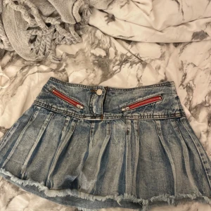 Kort jeanskjol från Chemilan med röda dragkedjor - Midjemåttet är 34cm. Säljer en cool kort jeanskjol från Chemilan med plisserad design och rå kant nedtill. Kjolen har två röda dragkedjor framtill som ger en edgy vibe. Tillverkad i 100% bomull och har klassisk blå tvättad denimlook. Perfekt för dig som gillar streetstyle.
