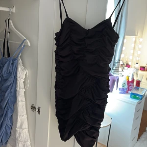 Svart ruched kort klänning - Supersnygg svart kort klänning med tunna axelband och ruched detaljer över hela klänningen. Sitter tight och har en dragkedja i sidan för enkel påklädning. Perfekt för dig som vill sticka ut med en cool och trendig look.