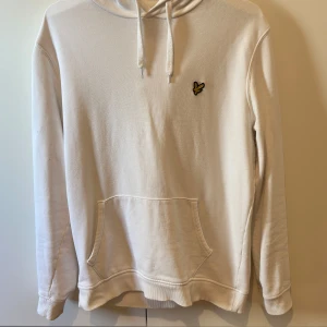 Vit hoodie från Lyle & Scott, storlek L - Vit hoodie från Lyle & Scott med klassisk gul logotyp på bröstet. Tröjan har huva med snörning, stor magficka och ribbade muddar. Perfekt för chill dagar och enkel att matcha med jeans eller joggers.