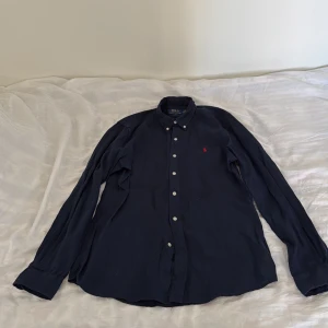 Mörkblå linneskjorta Polo Ralph Lauren - Mörkblå skjorta från Polo Ralph Lauren i slim fit med klassisk krage, vita knappar och röd broderad logga på bröstet. Tillverkad i svalt linne och har långa ärmar. Perfekt för en clean och stilren look.
