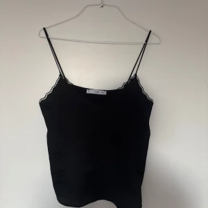 Svart linnetopp med spets från Mango - Svart linnetopp från Mango med tunna axelband och vacker spetskant längs urringningen. Toppen har en stilren och enkel design som passar perfekt till många olika looks. Materialet känns lätt och svalt, och den har en feminin v-ringad form.