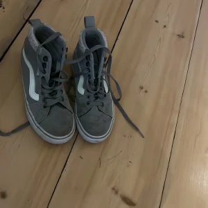 Grå Vans höga sneakers med foder - Säljer ett par grå höga Vans sneakers med vit sula och klassisk vit sidostrip. Skorna har snörning och är fodrade med mjukt material för extra värme. Perfekta för dig som vill ha en streetig look även under kallare dagar.