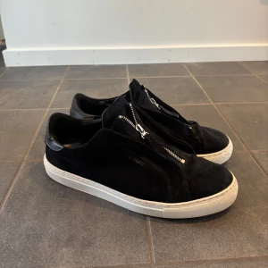 Svarta sneakers från Axel Arigato - Svarta sneakers i mocka från Axel Arigato med vit platt sula och dragkedja framtill. Snygg minimalistisk design med diskret logga i guld på sidan. Det är inga slags defekter på skorna. De är ganska använda då skicket är på ungefär 6/10.