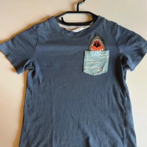 Blå t-shirt med hajficka från H&M - Blå t-shirt från H&M i bomull med korta ärmar. På bröstet finns en cool ficka med vågmönster och en haj som sticker upp, vilket ger en lekfull och unik look. Perfekt för barn som gillar havet och djurmotiv.