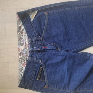 Replay blå jeans med blommigt foder - Snygga blå jeans från Replay med raka ben och coolt blommigt innerfoder. Klassisk femficksmodell, detaljerade sömmar och svart Replay-märke med paljetter bak i midjan. Perfekta för dig som vill ha något unikt och stilrent i garderoben. Stl 28/34