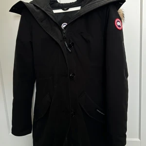 Svart vinterjacka  Canada Goose - Säljer en svart vinter jacka från Canada Goose med huva och snygg pälskant. Jackan har klassisk logga på ärmen, stora fickor och stängs med både dragkedja och knappar. Perfekt för kalla vinterdagar och riktigt stilren look. Modell rossclair endast använd en vinter, inga defekter eller skador!