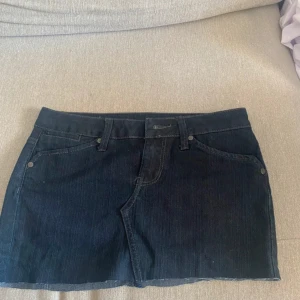 Mörk jeanskjol med broderade bakfickor - Snygg mörkblå jeanskjol i kort modell med klassiska fickor fram och bak. Bakfickorna har coola broderade vågmönster med nitar som ger extra edge. Kjolen har hällor för bälte och rå kant nedtill för en trendig look.