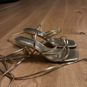 Guldiga klackar med snörning från Even&Odd - Snygga guldiga klackar från Even&Odd med tunna remmar och snörning som kan lindas runt vristen. Skorna har fyrkantig tå och låg klack, perfekta för att lyfta din outfit. BARA ANVÄNDA EN GÅNG FÖR BAL