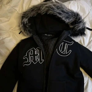 Svart pufferjacka med fuskpäls och brodyr - Svart pufferjacka med stor huva kantad av gråsvart fuskpäls. Jackan har quiltat innerfoder, stora broderade bokstäver på bröstet och en diskret svart örn på ryggen. Perfekt för kalla dagar och riktigt snygg streetstil.