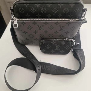 Svart axelväska med monogram - Snygg svart axelväska med klassiskt monogrammönster och silverfärgade detaljer. Väskan har bred justerbar axelrem med logga och en liten avtagbar ficka framtill. Tillverkad i skinn och levereras med dustbag.