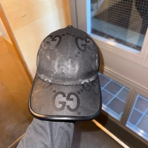 Svart Jumbo Gucci keps - Snygg svart keps från Gucci med ikoniskt GG-mönster över hela kepsen. Kepsen har böjd skärm med svart kant och justerbar rem baktill. Tillverkad i slitstarkt tyg med exklusiv känsla, perfekt för dig som vill sticka ut med en lyxig accessoar.