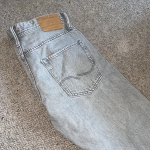 Ljusgrå jeans från Jack & Jones - Säljer ett par ljusgrå jeans från Jack & Jones med klassisk femficksdesign och normal passform. Jeansen har bälteshällor, knappgylf och är tillverkade i mjukt denimtyg. Perfekta för en avslappnad och stilren look.