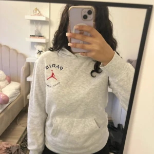 Grå Paris Saint Germain hoodie - Säljer en ljusgrå hoodie från Jordan x Paris Saint Germain med röd logga och text på bröstet samt stor Paris 23-tryck på ryggen. Tröjan har huva, magficka och är långärmad. Perfekt för dig som gillar sportig streetstyle.