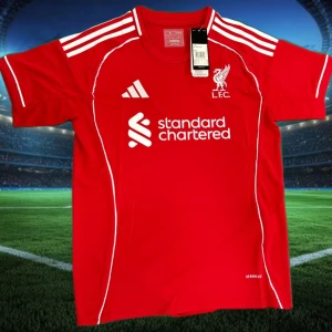 Liverpool röd fotbollströja Adidas - Riktigt snygg röd Liverpool fotbollströja från Adidas med vita detaljer och klubbmärke på bröstet. Klassiska tre ränder på axlarna och tryckt sponsorlogga framtill. Tillverkad i lätt och ventilerande material, perfekt för match eller träning.