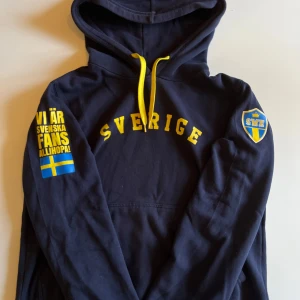 Sverige hoodie Supreme Basics L - Mörkblå hoodie med gula detaljer och tryck 'Sverige' på bröstet. Gult snöre i huvan, tryck på ena ärmen och svensk flagga. Perfekt för dig som vill visa ditt stöd för svenska landslaget. Mjuk och skön bomullskänsla.