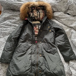 Dunjacka från Burberry Brit storlek medium - Snygg grön/grå dunjacka från Burberry Brit med klassiskt rutigt foder, stor huva med pälskrage och detaljer i guld. Jackan har dragkedja, knappar och praktiska fickor. Perfekt för kalla dagar och riktigt stilren look. Storlek large men är väldigt liten i storlek så mer än mindre small eller vanlig medium. I bra skick men behöver kemtvättas. Äkta!