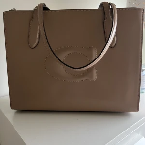 Beige handväska från Coach i skinn - Stilren beige handväska från Coach i slätt skinn med präglad C-logga på framsidan. Väskan har två smala handtag, dragkedja upptill och en klassisk tag med Coach-logga. Perfekt för dig som gillar minimalistisk och lyxig design.