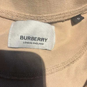 Beige Burberry pikétröja med logga - Snygg beige pikétröja från Burberry med klassisk krage, korta ärmar och broderad TB-logga på bröstet. Ikoniskt rutmönster på axlarna ger en extra touch. Tillverkad i bomull för en skön känsla.