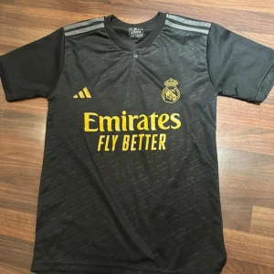 Real Madrid Vini Jr. svart matchtröja - Snygg svart Real Madrid fotbollströja från Adidas med Vini Jr. och nummer 7 på ryggen. Tröjan har gula tryck, klubbmärke på bröstet och klassiska Adidas-ränder på axlarna. Tillverkad i lätt och ventilerande polyester, perfekt för träning eller match.