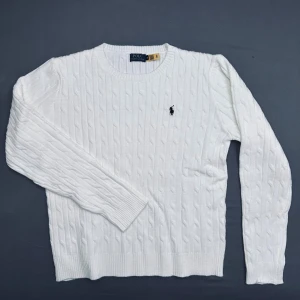 Vit kabelstickad tröja från Polo Ralph Lauren - Vit kabelstickad tröja från Polo Ralph Lauren med klassisk rund hals och broderad marinblå logga på bröstet. Tröjan har ribbade muddar vid ärmslut och nederkant, samt långärmad passform. Perfekt för dig som gillar stilrena och tidlösa plagg.