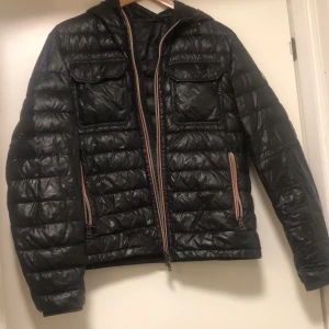 Svart dunjacka från Moncler - Säljer en svart dunjacka från Moncler med quiltad design och huva. Jackan har två bröstfickor med lock, sidofickor med dragkedja och dubbla dragkedjor framtill. Materialet är glansigt och jackan är perfekt för kalla dagar. Den har defekter se bild 4 och 5. Det finns även småmål längst ner på ärmarna, men inget av detta märks när man har på sig jackan.
