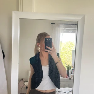 Svart stickad väst med bred krage - Säljer en svart stickad väst med bred krage och öppen front. Västen har en mjuk och fluffig yta som ger en cool look. Passformen är normal och modellen är ärmlös, vilket gör den enkel att styla över en t-shirt eller topp.