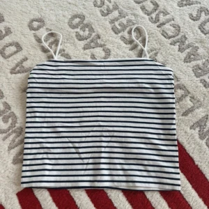 Randig linnetopp från Gina Tricot - Säljer en vit och marinblå randig linnetopp från Gina Tricot. Toppen har tunna axelband och är i mjuk bomull, perfekt för varma dagar. Enkel och stilren design som är lätt att matcha med jeans eller kjol.