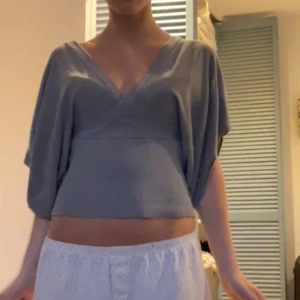 Grå omlottopp med vida ärmar - Säljer en grå omlottopp med v-ringning och vida, korta ärmar. Toppen är croppad och har en snygg omlottdesign som ger en cool och avslappnad vibe. Perfekt att styla med höga jeans eller kjol.