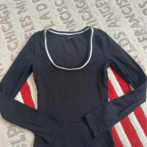 Svart långärmad topp Gina Tricot - Svart långärmad topp från Gina Tricot med vit kant runt den djupa, rundade urringningen. Topp i mjukt material som sitter snyggt och passar perfekt till jeans eller kjol. Enkel men ändå med en cool detalj vid halsen.