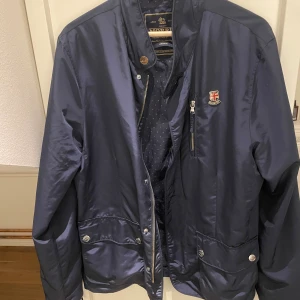 Mörkblå vindjacka från Morris, M - Snygg mörkblå vindjacka från Morris med klassisk design. Jackan har dragkedja, knappar, flera fickor och ett litet emblem på bröstet. Tillverkad i glansig polyester och har långärmad passform. Perfekt för dig som gillar stilrena och tidlösa plagg.